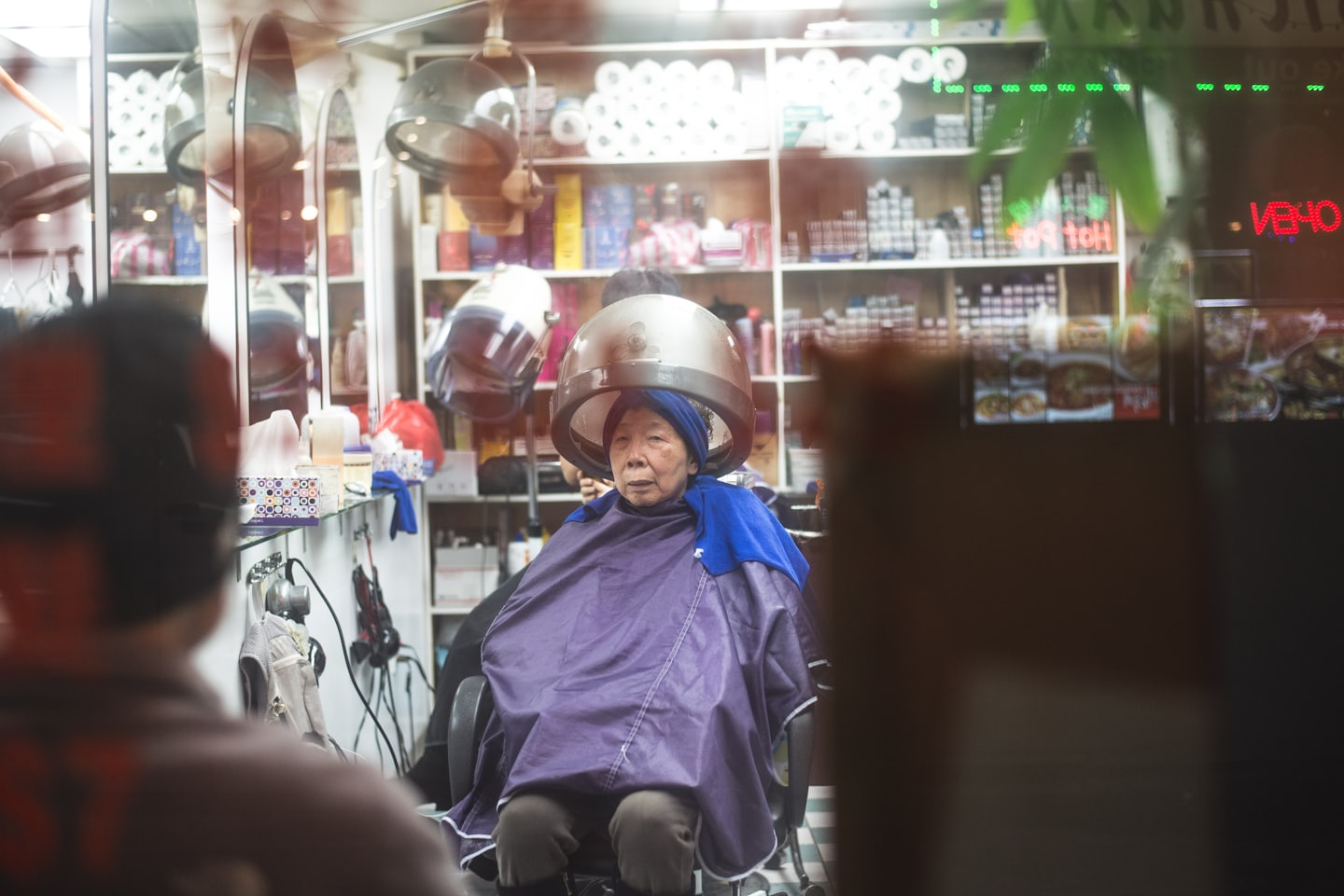 Kunde unter einer Trockenhaube in einem Friseursalon mit Regalen voller Lockenwickler.